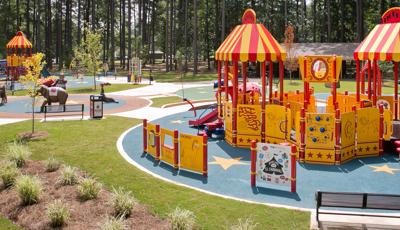 In the foreground yellow and red colored circus themed play structure has roof panels designed like circus tents. A second circus themed play structure sits in the background along with a surrounding wall of tall trees.