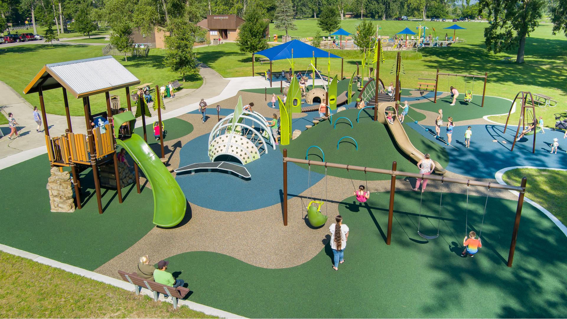 Elevated view of a nature themed playground with climbable incorporated hillsides, large trees fill the background with a large lake beyond that.