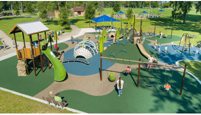 Elevated view of a nature themed playground with climbable incorporated hillsides, large trees fill the background with a large lake beyond that.