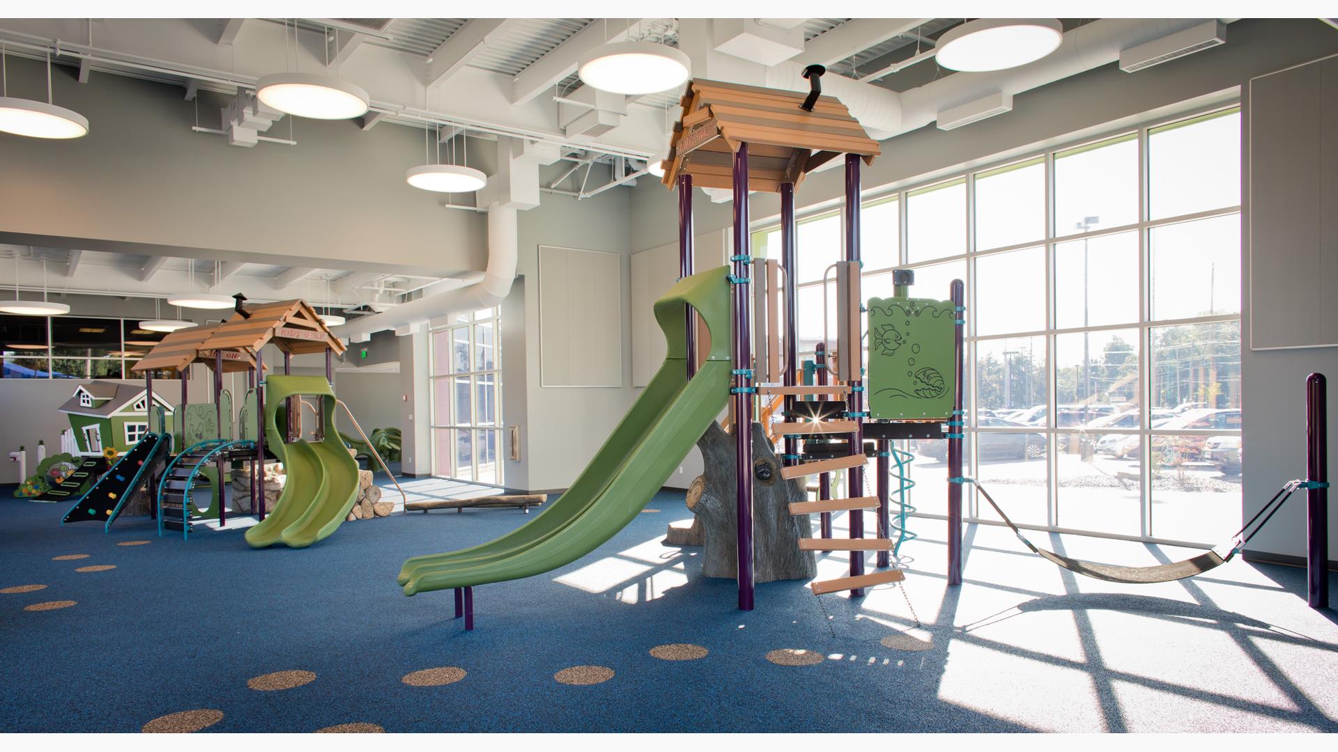 Indoor play area at St. Vincent Children's hospital with sun coming through ceiling to floor windows. There are three playgrounds with nature-inspired theme and colors with blue rubber surfacing. 