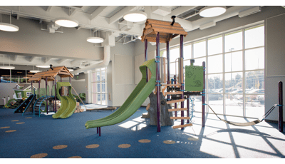 Indoor play area at St. Vincent Children's hospital with sun coming through ceiling to floor windows. There are three playgrounds with nature-inspired theme and colors with blue rubber surfacing. 