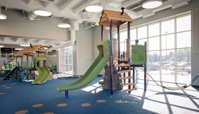 Indoor play area at St. Vincent Children's hospital with sun coming through ceiling to floor windows. There are three playgrounds with nature-inspired theme and colors with blue rubber surfacing. 