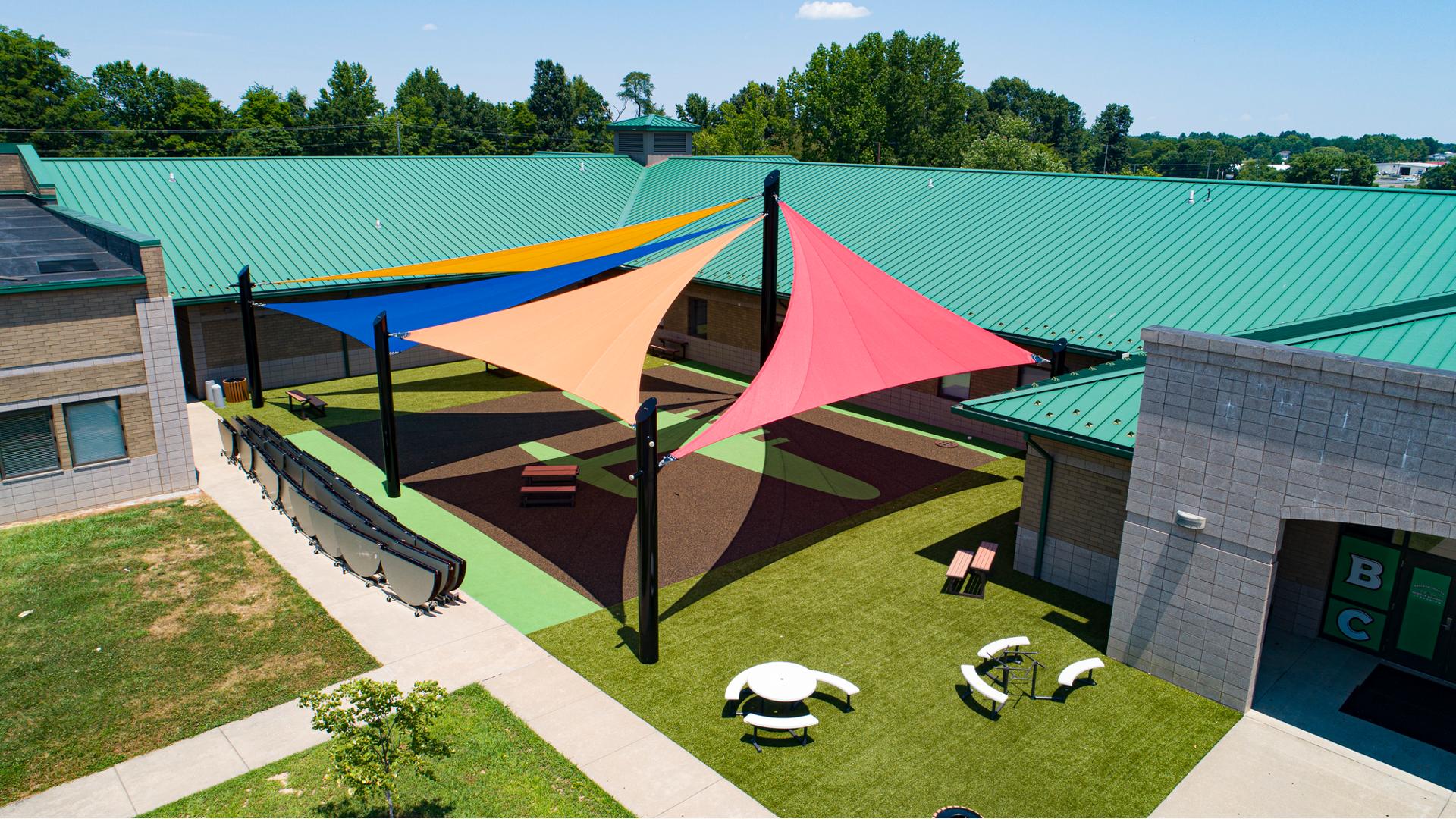 A large open area nestled into a building with an airplane silhouette designed into the turf. Four large triangular shade sales connect to four different posts over the open turfed area. A line of folded circular tables line a side walk next to the turfed area.