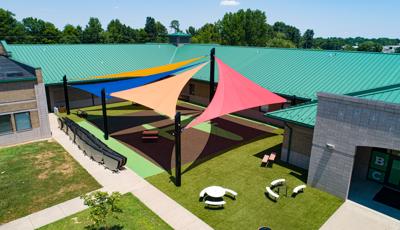 A large open area nestled into a building with an airplane silhouette designed into the turf. Four large triangular shade sales connect to four different posts over the open turfed area. A line of folded circular tables line a side walk next to the turfed area.