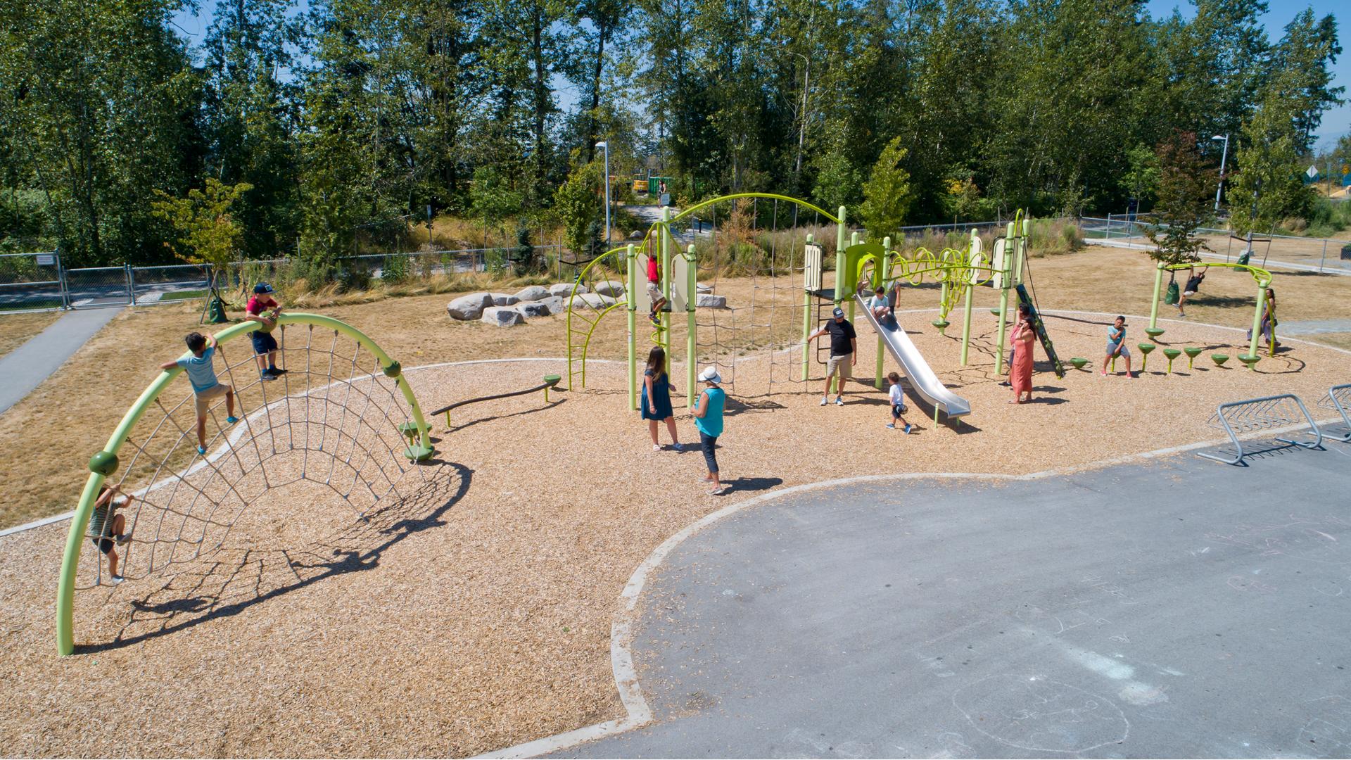 Families play at an outdoor park with a full tree line in the background. Children climb on a half-moon arch play structure with ropes like a spider’s web. Other families play on a second play structure with climbers and slides.