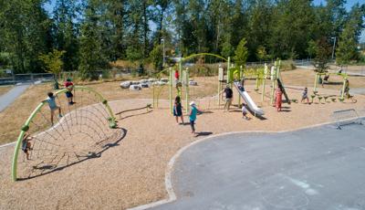 Families play at an outdoor park with a full tree line in the background. Children climb on a half-moon arch play structure with ropes like a spider’s web. Other families play on a second play structure with climbers and slides.