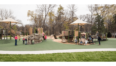 On an overcast day children play on the nature-inspired PlayBooster playground while two moms watch. A narrow tree-line creates a natural border around the playground.