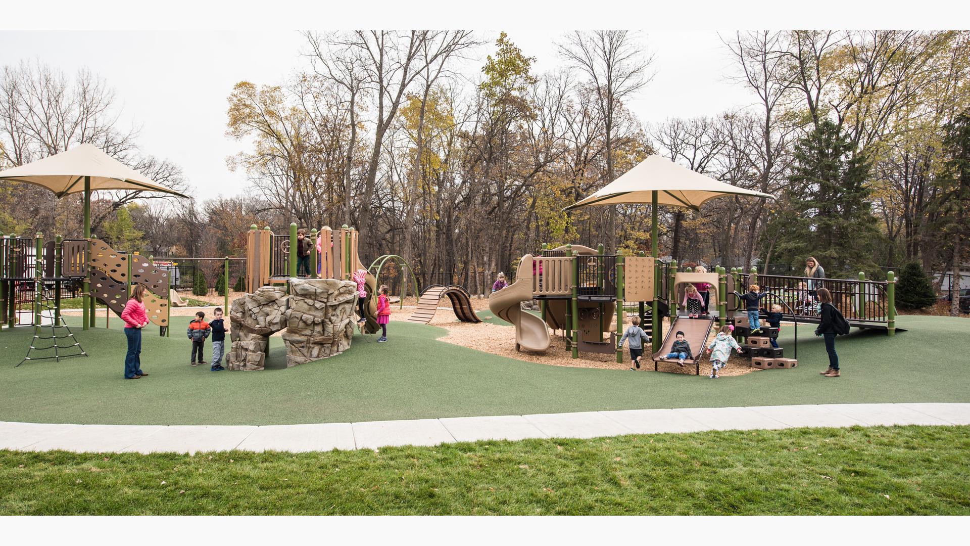 On an overcast day children play on the nature-inspired PlayBooster playground while two moms watch. A narrow tree-line creates a natural border around the playground.
