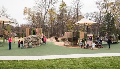 On an overcast day children play on the nature-inspired PlayBooster playground while two moms watch. A narrow tree-line creates a natural border around the playground.