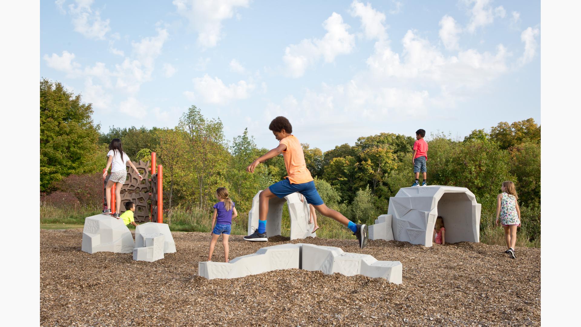 Boy in orange shirt leaps from "rock," while other children stand on "rock formations." A freestanding climber stands in the background.