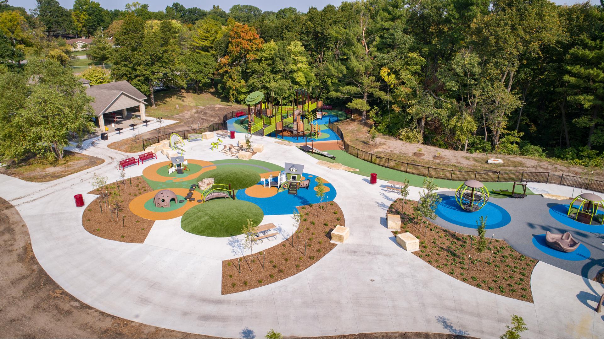 Aerial image of playground site which includes several play areas for kids of all ages. For smaller kids there are two play house themed playstructures. And for older kids there is a nature themed playground with accessible ramps. 