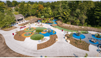 Aerial image of playground site which includes several play areas for kids of all ages. For smaller kids there are two play house themed playstructures. And for older kids there is a nature themed playground with accessible ramps. 