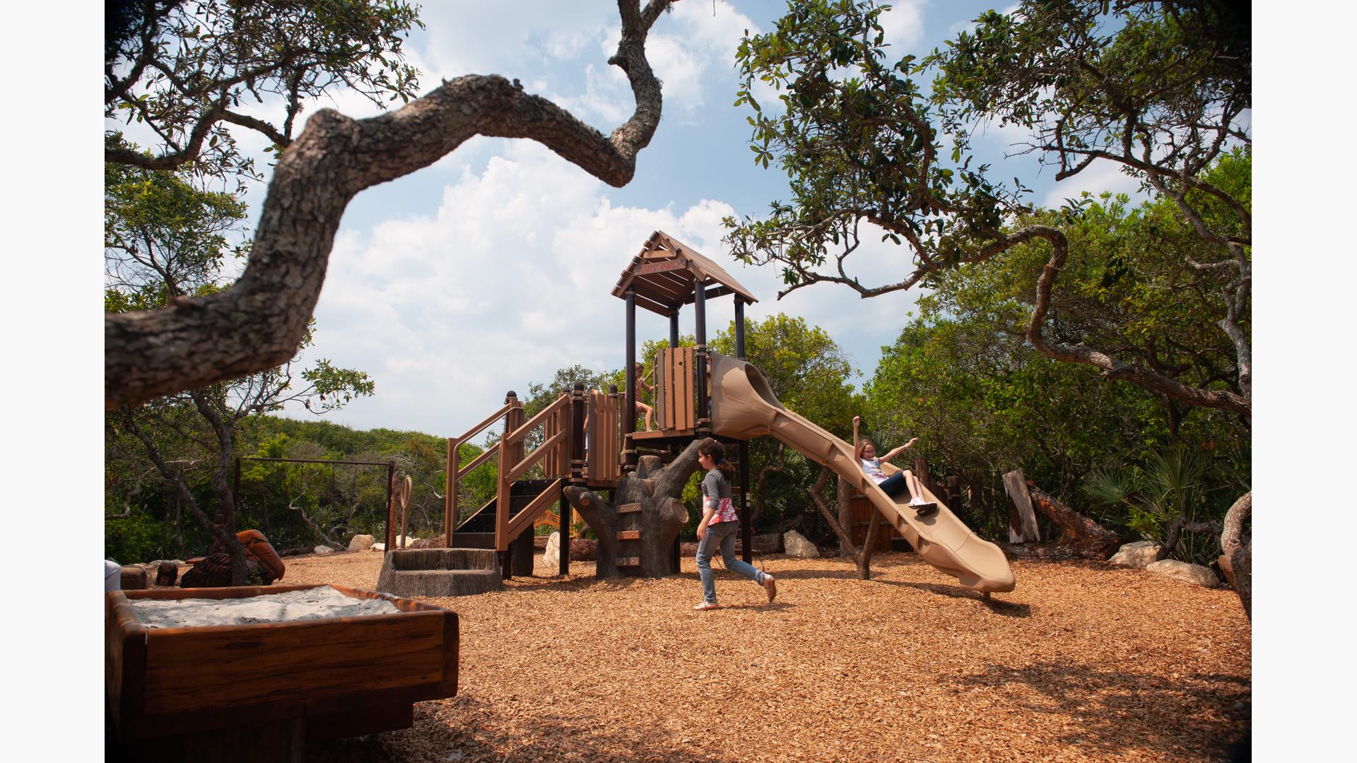 Girl riding down slide as friend in grey crosses playground. Play-naturally themed, Ponce Preserve sits in the natural lay of the of the Florida landscape.