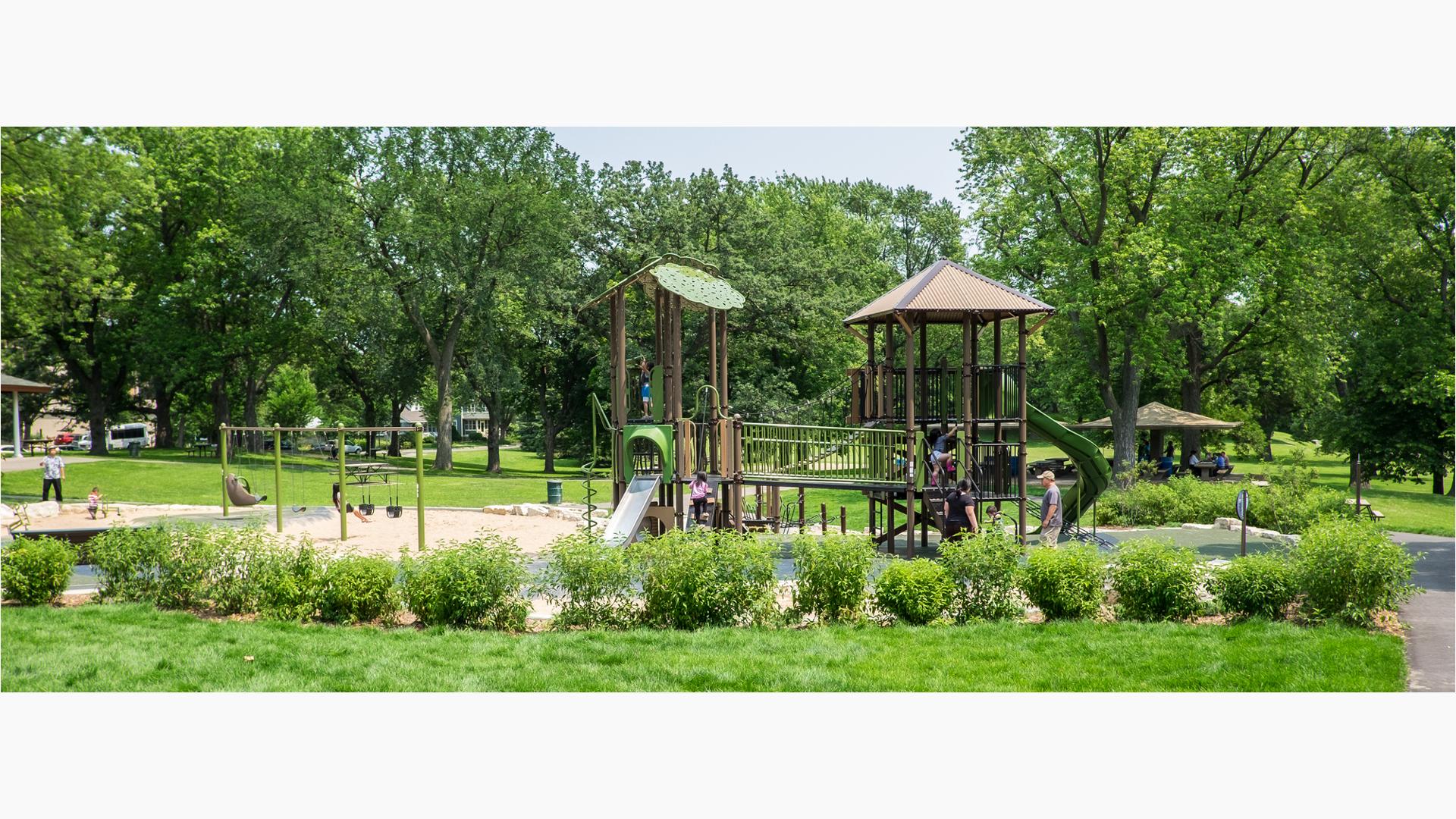 Nature-inspired playground in a heavily wooded park. Children sit on the double swing set. Parents watch from the outskirts. More children play on the main PlayBooster play structure.