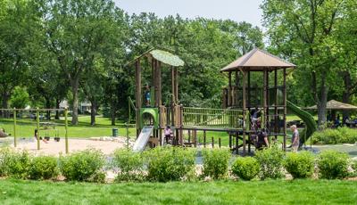 Nature-inspired playground in a heavily wooded park. Children sit on the double swing set. Parents watch from the outskirts. More children play on the main PlayBooster play structure.