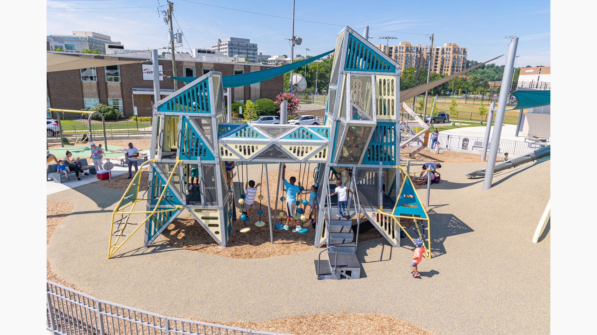 Children play on a playground of two towers with an elevated connecting crawl tunnel in an urban setting. Families sit under a large shade sail in the background to the left.