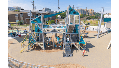 Children play on a playground of two towers with an elevated connecting crawl tunnel in an urban setting. Families sit under a large shade sail in the background to the left.