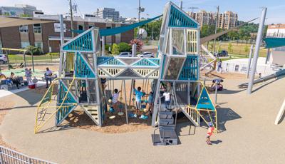 Children play on a playground of two towers with an elevated connecting crawl tunnel in an urban setting. Families sit under a large shade sail in the background to the left.