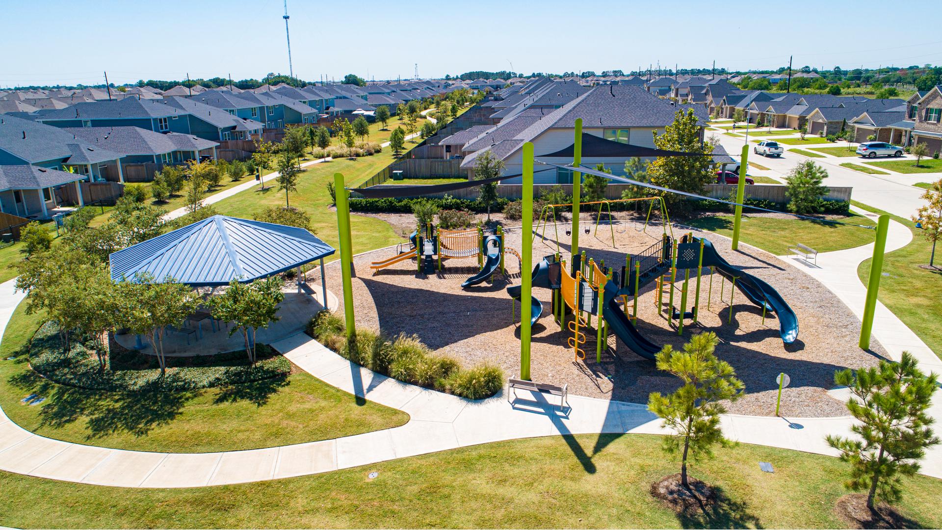 A park set in a neighborhood development has large triangular navy sail shades above two separate play structures with a pavilion nearby with picnic tables.
