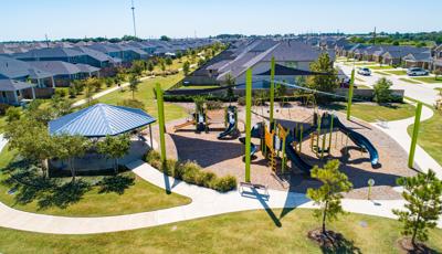 A park set in a neighborhood development has large triangular navy sail shades above two separate play structures with a pavilion nearby with picnic tables.