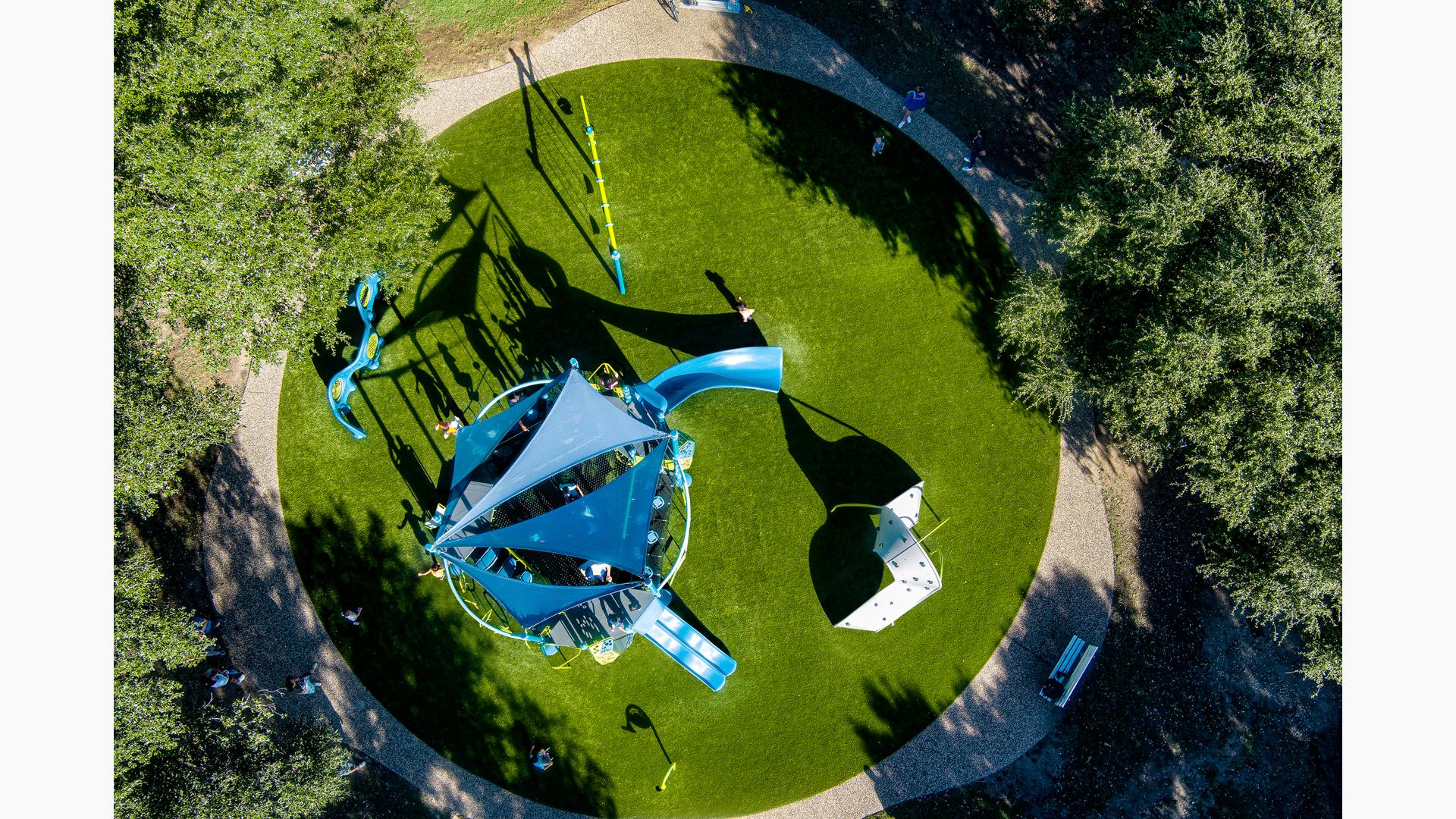 Bird's eye view of a circular park play area with a main play structure with shade sails and other surrounded climbers, swings, and panel play activities.