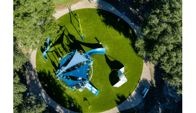 Bird's eye view of a circular park play area with a main play structure with shade sails and other surrounded climbers, swings, and panel play activities.