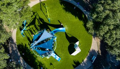Bird's eye view of a circular park play area with a main play structure with shade sails and other surrounded climbers, swings, and panel play activities.