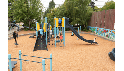 Whittier Elementary School playground sits 
 tucked away in a local neighborhood. A boy in blue shirt climbs rock wall, while another climbs net climber. A little boy sits at the bottom of slide.