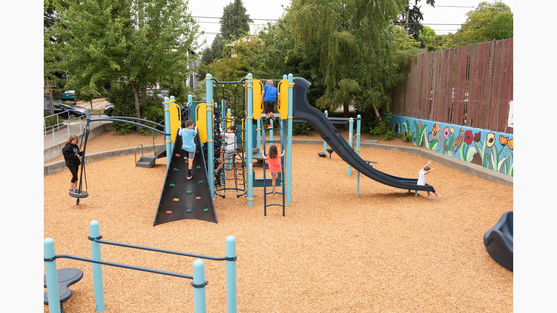 Whittier Elementary School playground sits 
 tucked away in a local neighborhood. A boy in blue shirt climbs rock wall, while another climbs net climber. A little boy sits at the bottom of slide.