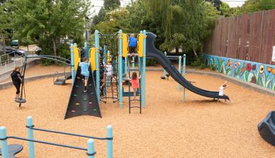 Whittier Elementary School playground sits 
 tucked away in a local neighborhood. A boy in blue shirt climbs rock wall, while another climbs net climber. A little boy sits at the bottom of slide.