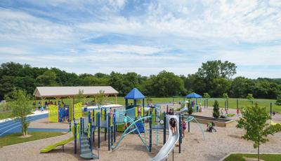 Children play on the colorful green and blue play structures with an additional standalone playground climber shaped as a large butterfly all surrounded by a blue running track.