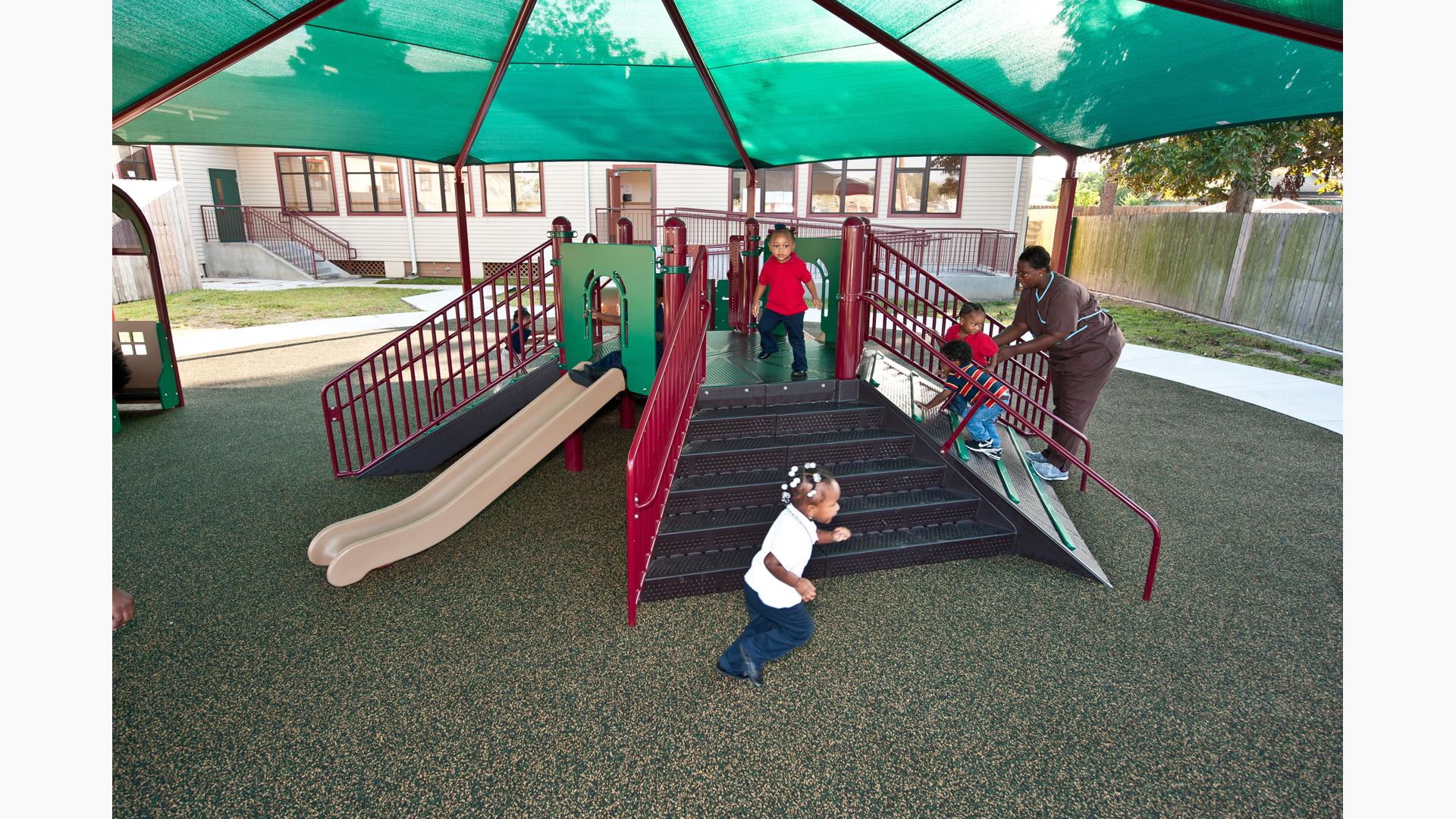 Woman in nursing attire help two boys climb up a ramped climber of a play structure under a large green shade. 