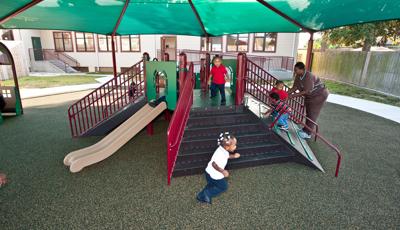 Woman in nursing attire help two boys climb up a ramped climber of a play structure under a large green shade. 