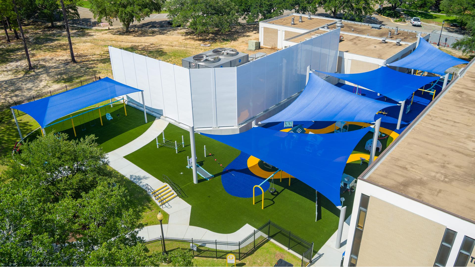 Elevated view of an inclusive playground nestled next to a building with large blue shade sails cover the play area.