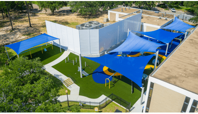 Elevated view of an inclusive playground nestled next to a building with large blue shade sails cover the play area.
