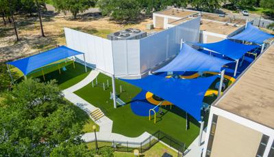 Elevated view of an inclusive playground nestled next to a building with large blue shade sails cover the play area.