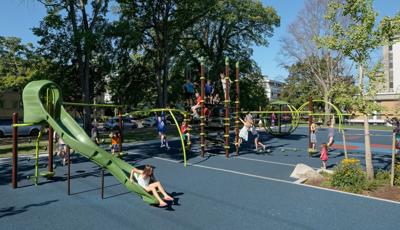 InVictoria Courthouse Playground, a girl is sitting at the bottom of slide . A group of kids swarm on the Netplex structure.