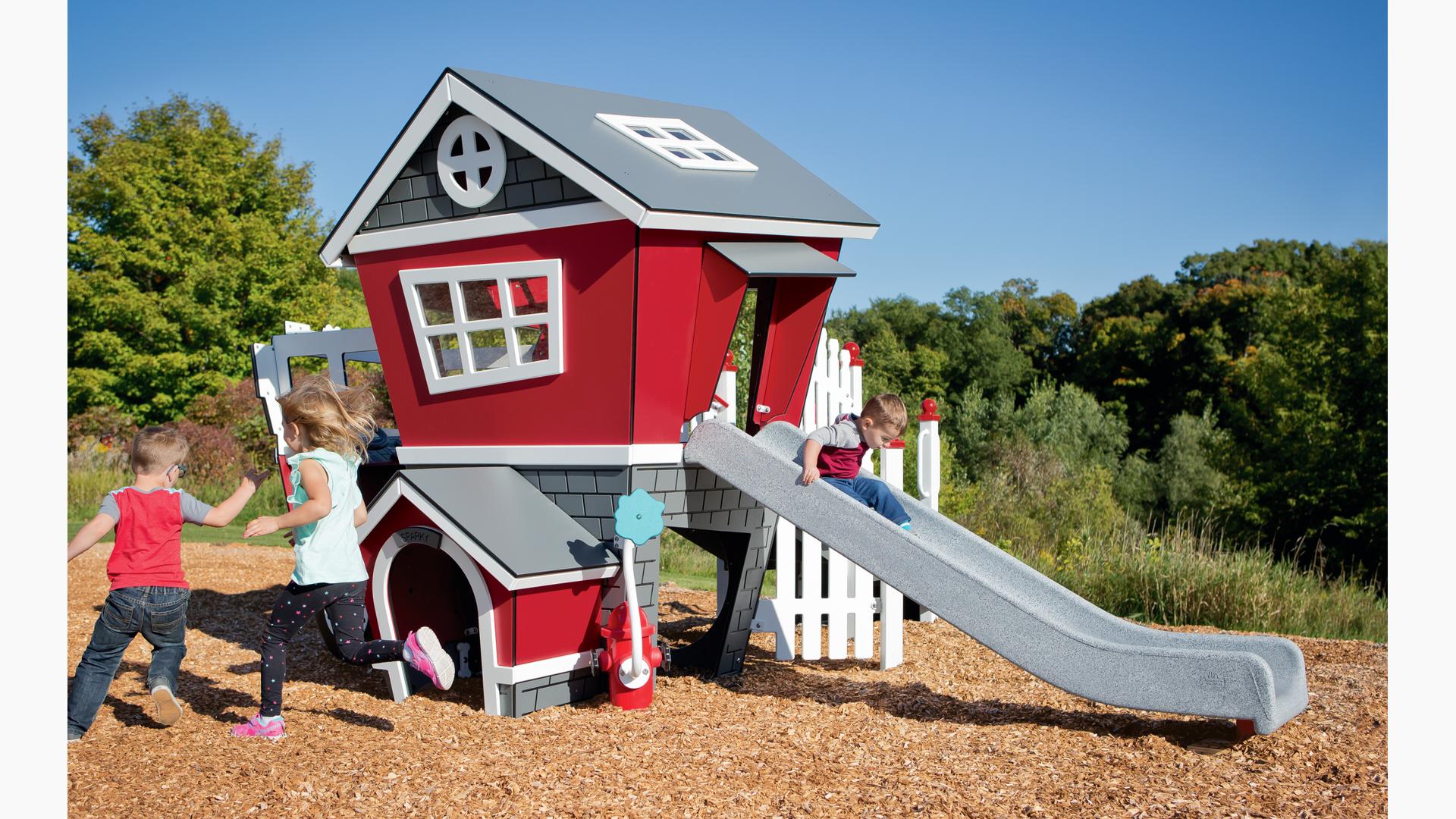 A boy and girl run around the  Smart Play Fire Station play structure together. Another boy rides down the Fire Station slide in the back.