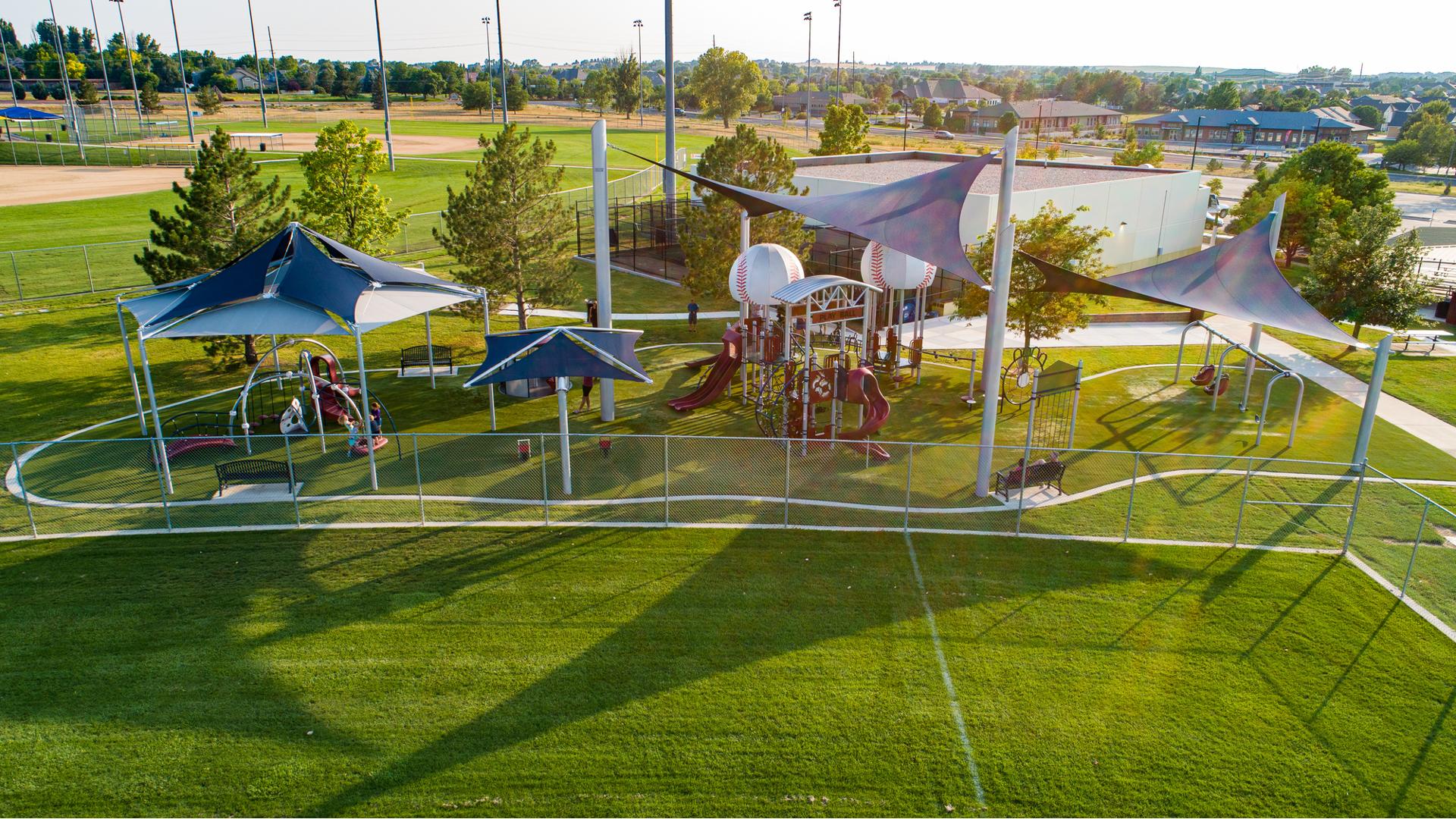 Large sports-themed playground set just beyond the fence of baseball fields outfields has custom baseball shaped roofs and large navy blue shade systems covering the play area.