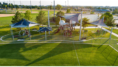 Large sports-themed playground set just beyond the fence of baseball fields outfields has custom baseball shaped roofs and large navy blue shade systems covering the play area.