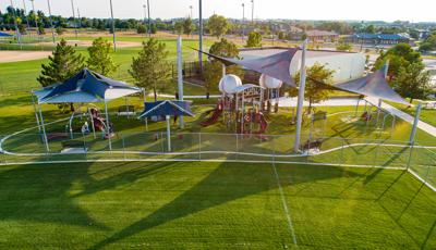 Large sports-themed playground set just beyond the fence of baseball fields outfields has custom baseball shaped roofs and large navy blue shade systems covering the play area.