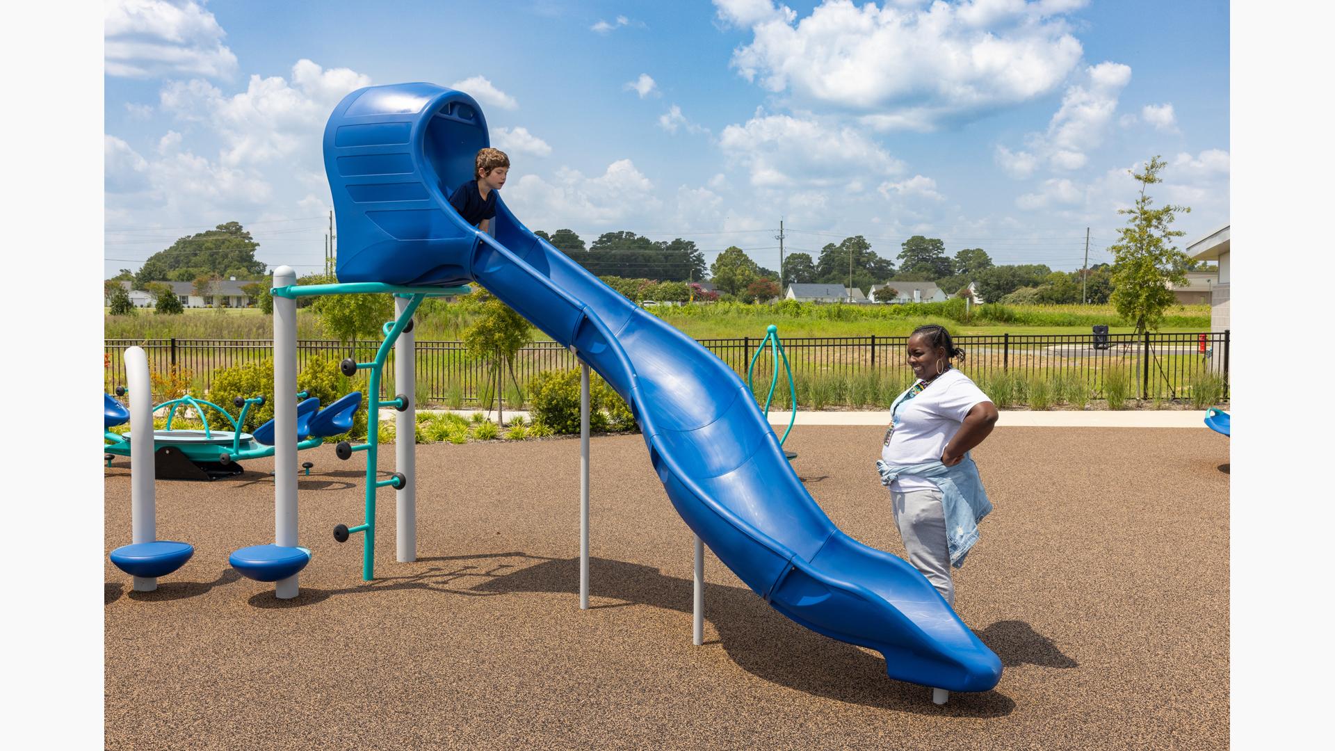 A boy prepares to slide down a standalone Rushwinder slide on a playground while a woman watches from below