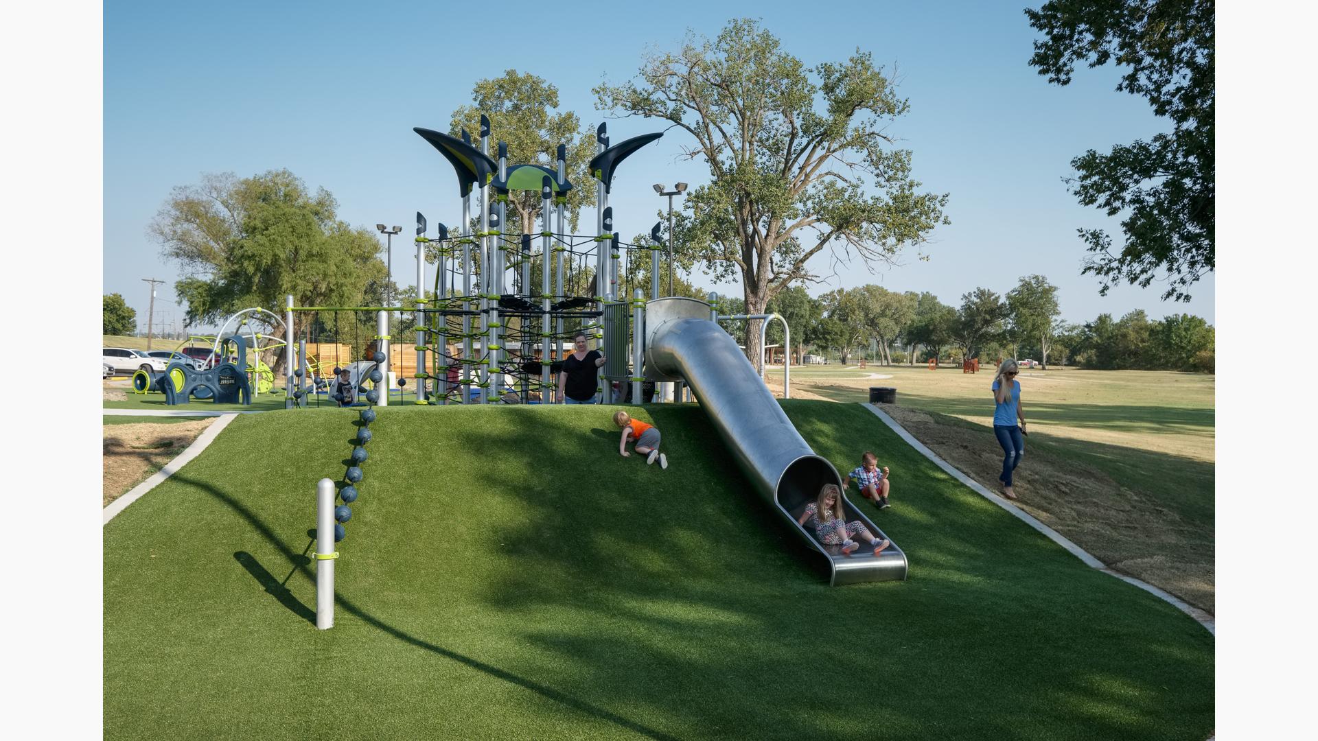 Children playing on a playground and going down a stainless tube slide with green artificial grass surfacing.