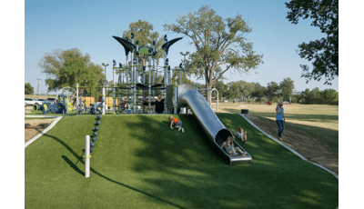 Children playing on a playground and going down a stainless tube slide with green artificial grass surfacing.