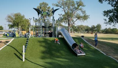 Children playing on a playground and going down a stainless tube slide with green artificial grass surfacing.