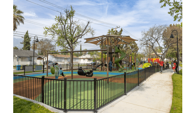 A black iron fence surrounds a outdoor play area with a large tower structures for larger kids and a smaller historical red street car designed play structure for younger children.