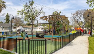 A black iron fence surrounds a outdoor play area with a large tower structures for larger kids and a smaller historical red street car designed play structure for younger children.
