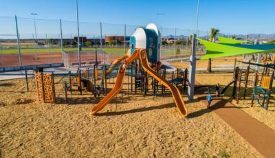 Kids playing on futuristic looking playground tower located in front of a baseball field.  There are two orange slides in the foreground with bright neon green and blue shade. 