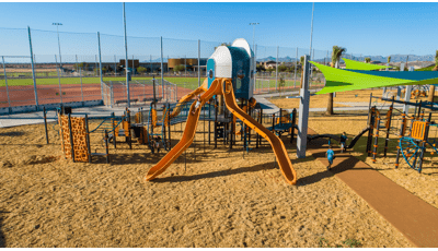 Kids playing on futuristic looking playground tower located in front of a baseball field.  There are two orange slides in the foreground with bright neon green and blue shade. 