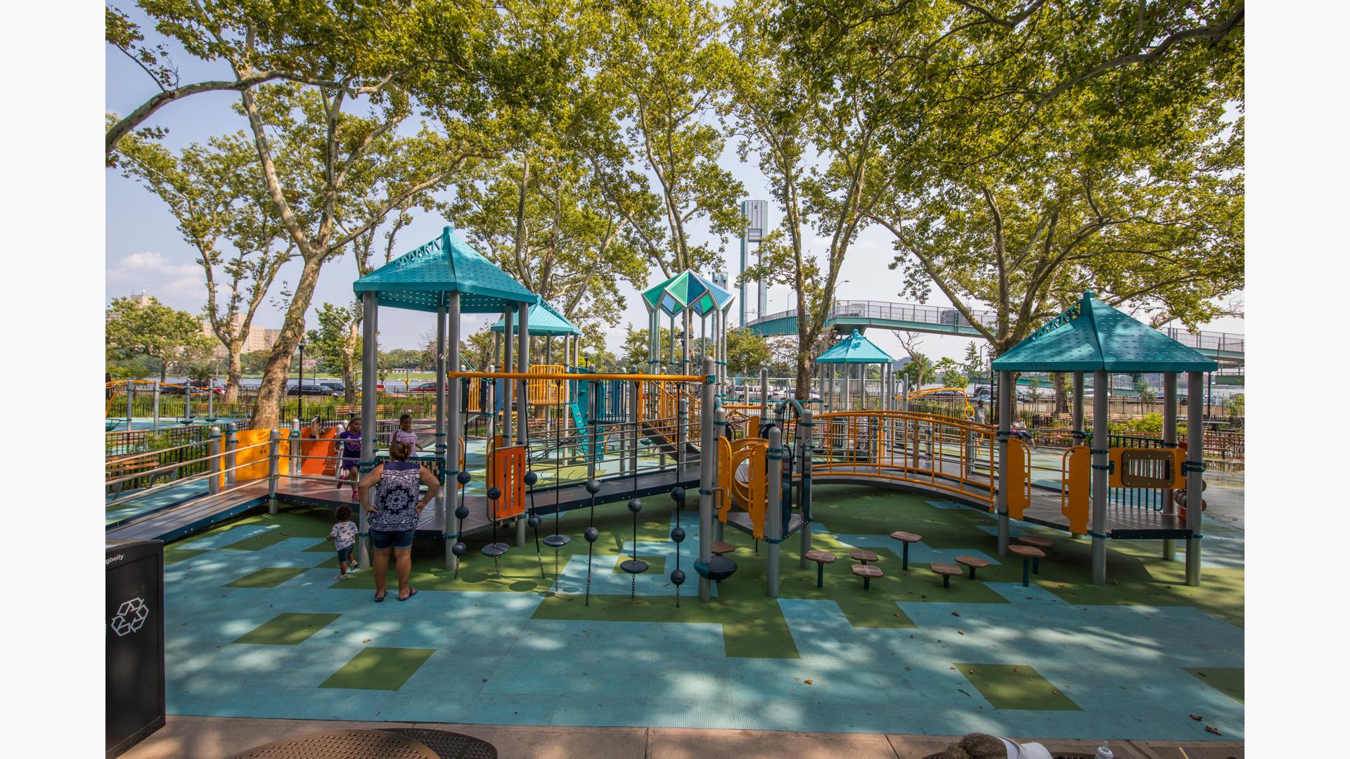 Children and family playing on a playground with light blue roofs.  The playground is shaded by tall green trees. 